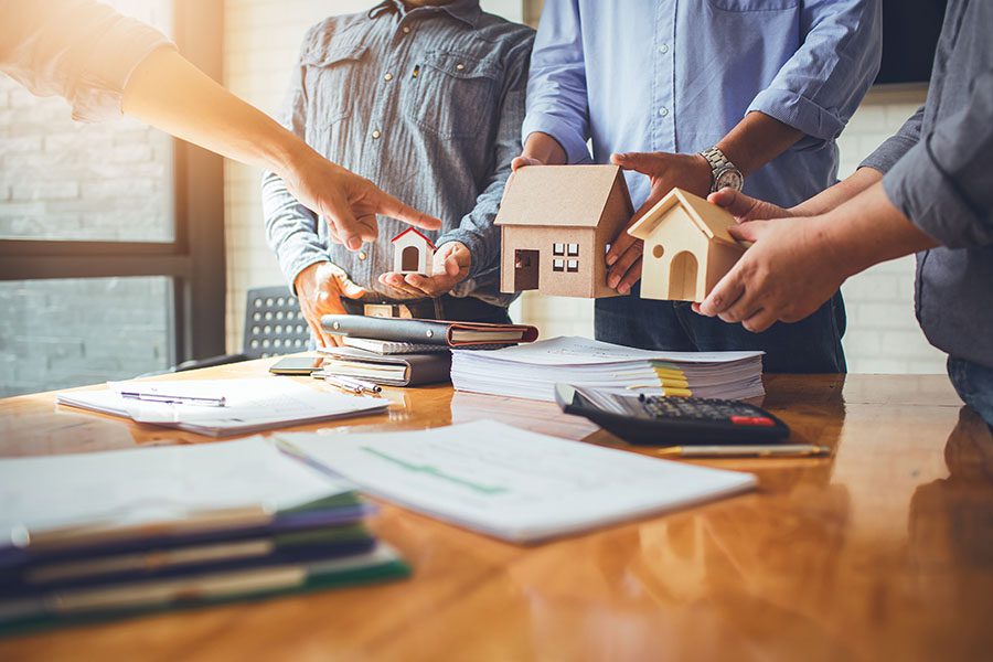 Insurance FAQs - Closeup View of Three Employees Standing in the Office Holding Up Different Wooden Figures of Homes for Boss to Pick Out