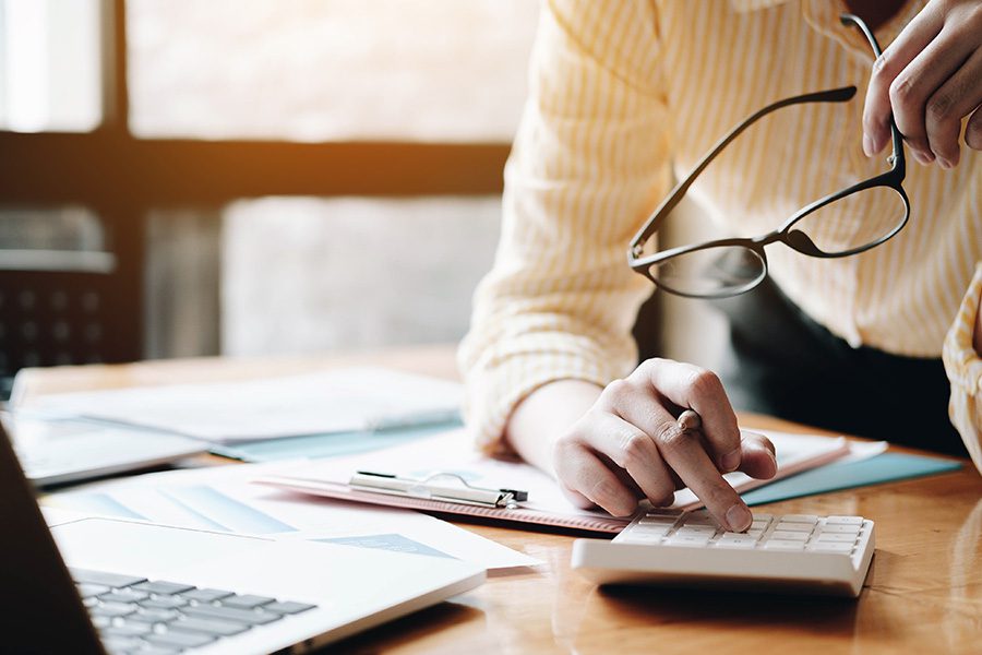 Tax Services - Close Up of Person in Yellow Shirt Typing on a Calculator While Holding Their Glasses and Looking at Their Open Laptop on a Desk Table