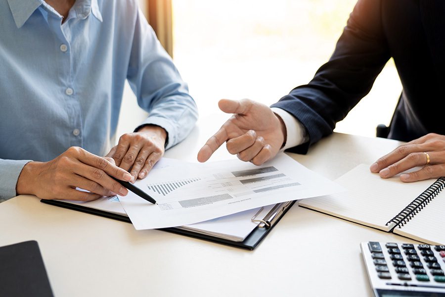 Wealth Management - Close Up of Professional Pointing at Graphs While Discussing with a Client at a White Desk with Sun Pouring In Through the Window Behind Them